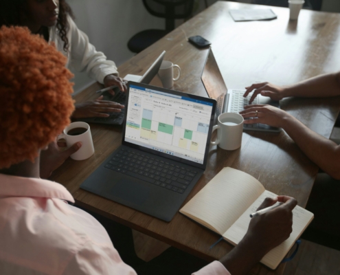 Three people in a meeting at a table discussing schedule on their Microsoft laptop